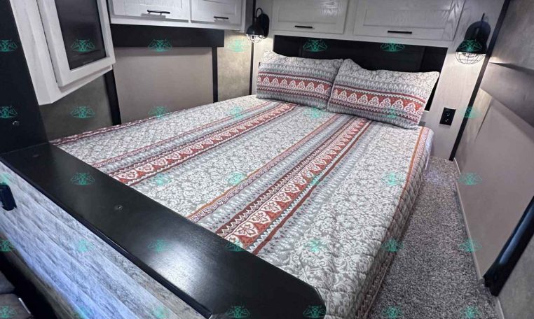 Interior of a travel trailer bedroom with a queen bed, patterned quilt, and overhead white cabinets above the headboard