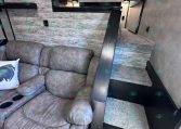 Interior of a modern trailer living area with a gray reclining sofa in the foreground, textured wood walls, and a short staircase leading to a compact kitchen with white cabinets and black trim under overhead lighting.
