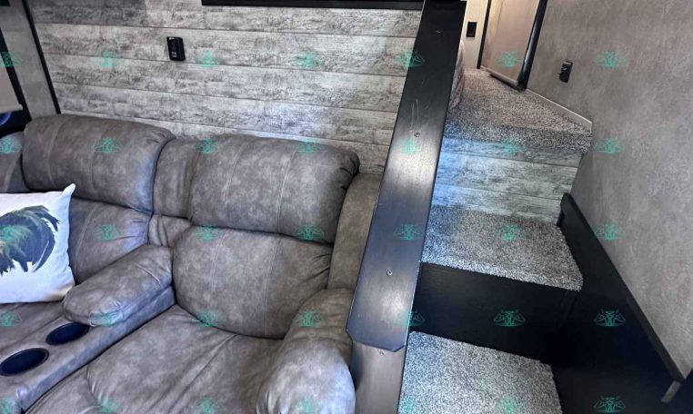 Interior of a modern trailer living area with a gray reclining sofa in the foreground, textured wood walls, and a short staircase leading to a compact kitchen with white cabinets and black trim under overhead lighting.