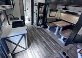 Interior of a modern travel trailer: kitchen with sink, microwave, and cabinets, adjacent dining table with benches, and a sofa in the foreground.