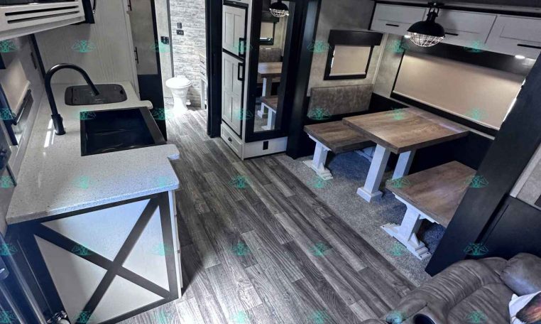 Interior of a modern travel trailer: kitchen with sink, microwave, and cabinets, adjacent dining table with benches, and a sofa in the foreground.