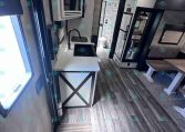 Interior of a modern travel trailer kitchen with sink, faucet, microwave, white cabinets, and a small dining area, plus a sofa and wood-like flooring.