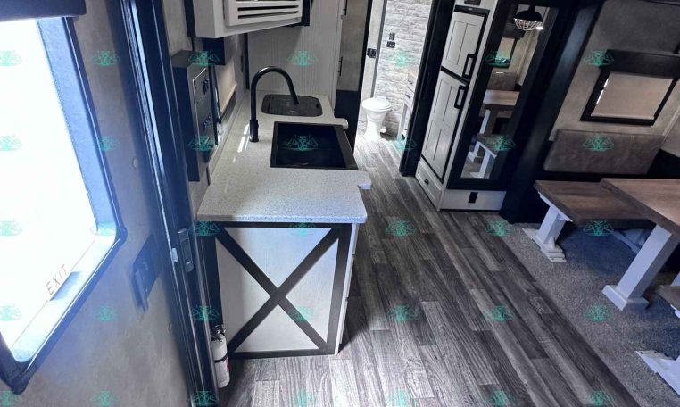 Interior of a modern travel trailer kitchen with sink, faucet, microwave, white cabinets, and a small dining area, plus a sofa and wood-like flooring.