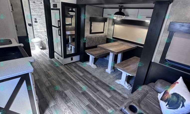 Cozy RV living area with a wooden dining table and bench seating, a sofa with a rooster pillow, and doorway to a bathroom.