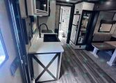 Interior of a modern RV kitchen and living area with sink, faucet, microwave, and refrigerator visible; dining booth and seating nearby.