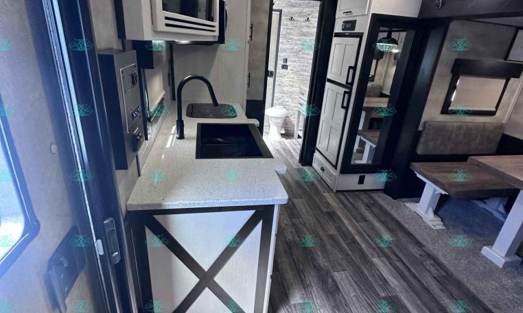 Interior of a modern RV kitchen and living area with sink, faucet, microwave, and refrigerator visible; dining booth and seating nearby.