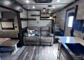 Interior of a modern trailer living area with a gray recliner sofa and a rooster-themed throw pillow, wood-look flooring, and a kitchenette island on the right.