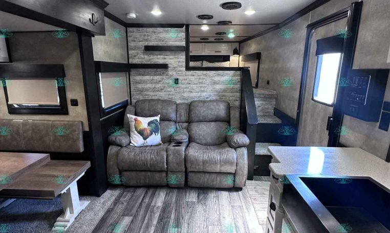 Interior of a modern trailer living area with a gray recliner sofa and a rooster-themed throw pillow, wood-look flooring, and a kitchenette island on the right.