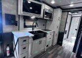 Modern RV kitchen: white cabinets, stainless steel microwave, and a black sink with a curved faucet; light gray subway backsplash and island counter.