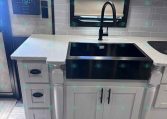 Compact kitchen area in a trailer with a black undermount sink, matte black faucet, light gray cabinets, and a gray tile backsplash across the counter edge.