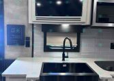 Compact kitchen in a trailer: white cabinetry, gray tiled backsplash, black induction cooktop, and a gooseneck faucet over a sink. A built-in microwave sits above the counter.