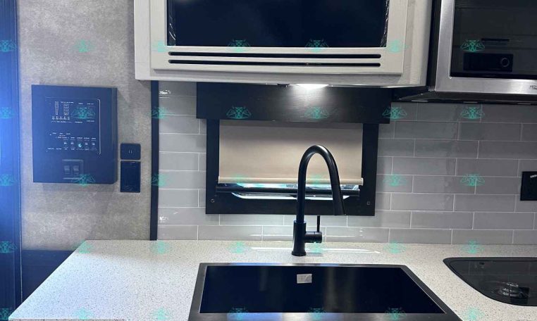 Compact kitchen in a trailer: white cabinetry, gray tiled backsplash, black induction cooktop, and a gooseneck faucet over a sink. A built-in microwave sits above the counter.