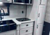 Modern kitchen in a light-gray trailer with white cabinets, stainless microwave, black sink, and a glass-top stove area.