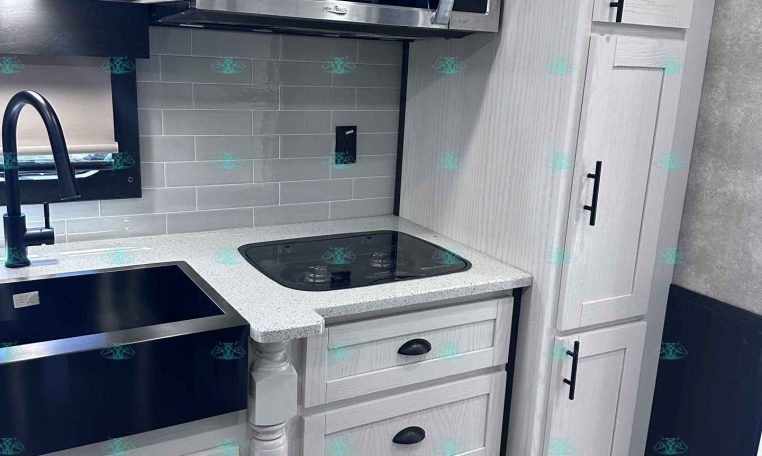 Modern kitchen in a light-gray trailer with white cabinets, stainless microwave, black sink, and a glass-top stove area.