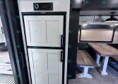 White paneled RV fridge with upper cabinet and Dometic control panel, next to a small dining table.