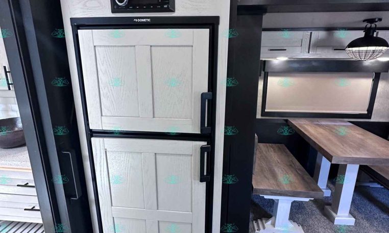 White paneled RV fridge with upper cabinet and Dometic control panel, next to a small dining table.