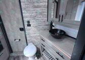 Modern trailer bathroom with gray wood panel walls, a black vessel sink, mirrored cabinet, countertop, and toilet.