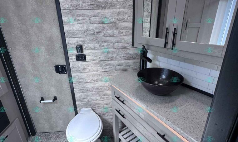 Modern trailer bathroom with gray wood panel walls, a black vessel sink, mirrored cabinet, countertop, and toilet.