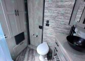 Small RV bathroom with wood-paneled walls, a white toilet, gray vanity with a black vessel sink, and a glass shower.