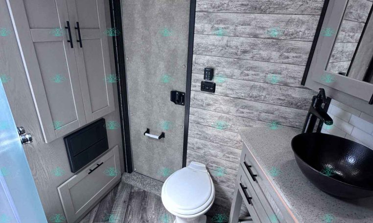 Small RV bathroom with wood-paneled walls, a white toilet, gray vanity with a black vessel sink, and a glass shower.