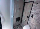 Compact RV bathroom with curved glass shower, gray cabinets, and a toilet against wood-paneled walls.