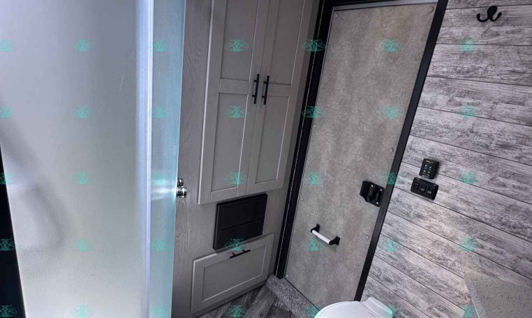 Compact RV bathroom with curved glass shower, gray cabinets, and a toilet against wood-paneled walls.