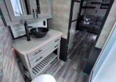 RV bathroom vanity with a black vessel sink, tall black faucet, gray cabinetry, and white tile backsplash; doorway to the living area with a gray sofa visible behind.