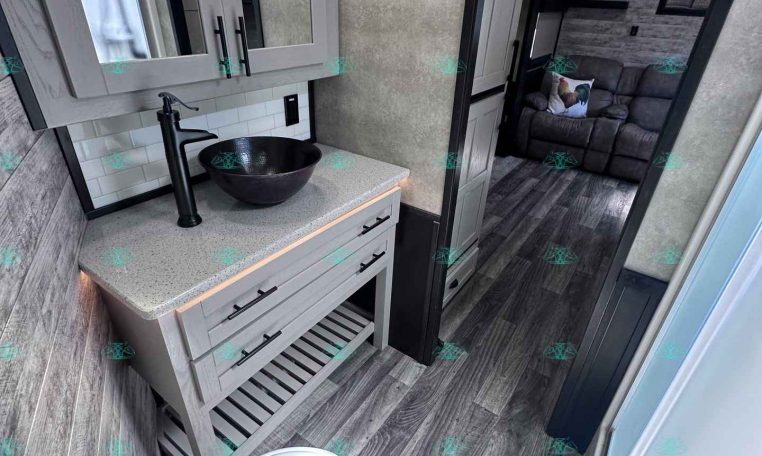 RV bathroom vanity with a black vessel sink, tall black faucet, gray cabinetry, and white tile backsplash; doorway to the living area with a gray sofa visible behind.