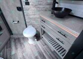 Small bathroom inside a trailer with a dark textured vessel sink on a speckled countertop, gray cabinetry, and a mirrored medicine cabinet above. The space has wood-look walls and a white toilet nearby.