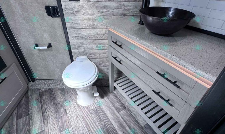 Small bathroom inside a trailer with a dark textured vessel sink on a speckled countertop, gray cabinetry, and a mirrored medicine cabinet above. The space has wood-look walls and a white toilet nearby.