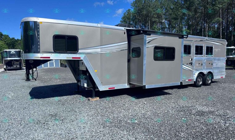Silver gooseneck horse trailer with living quarters parked on a gravel lot, trees in the background.