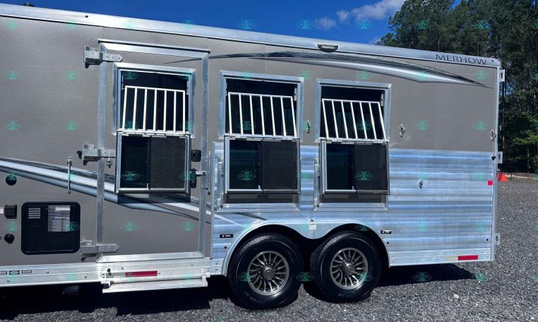 Silver Merhow aluminum horse trailer with three barred stall windows and chrome trim, parked on gravel under a blue sky.