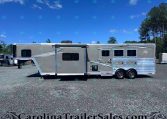 Side view of a long Merhow horse trailer parked on a gravel lot under a bright blue sky with trees in the background, ramp extended on the left.