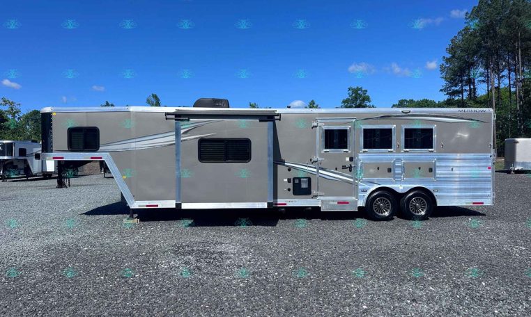 Side view of a long Merhow horse trailer parked on a gravel lot under a bright blue sky with trees in the background, ramp extended on the left.