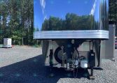 shiny aluminum trailer in a gravel lot, reflecting the surrounding trees and blue sky; hitch and support legs visible at the front