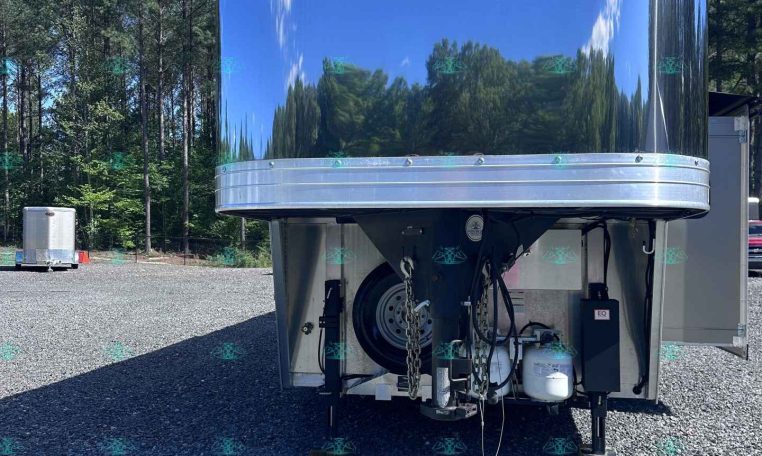 shiny aluminum trailer in a gravel lot, reflecting the surrounding trees and blue sky; hitch and support legs visible at the front