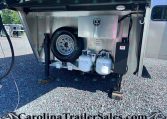Open trailer storage compartment with a spare tire, propane tanks, and a metal toolbox inside the bay on a gravel lot.
