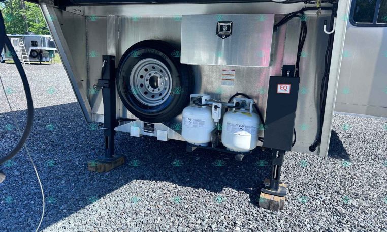 Open trailer storage compartment with a spare tire, propane tanks, and a metal toolbox inside the bay on a gravel lot.