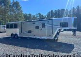 Silver travel trailer parked on a gravel lot with tall pine trees and a bright blue sky in the background, front-right stairs folded.