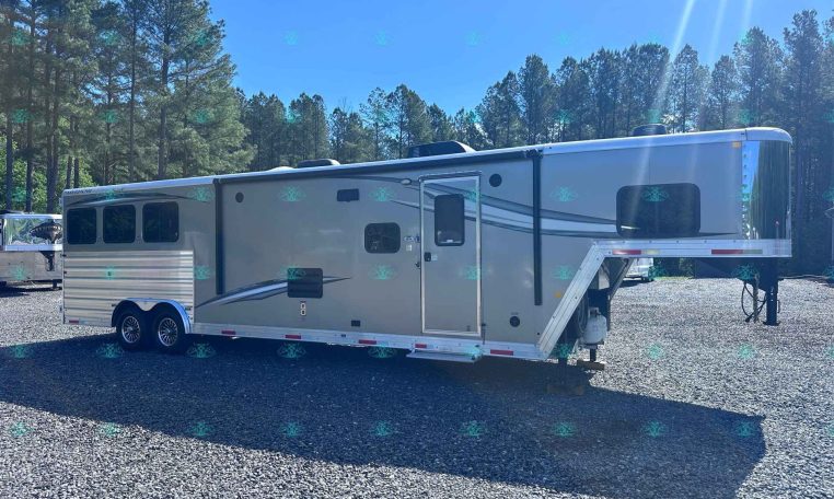 Silver travel trailer parked on a gravel lot with tall pine trees and a bright blue sky in the background, front-right stairs folded.