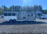 Side view of a long white and gray Merhow horse trailer parked on a gravel lot with tall pine trees and a blue sky behind it.