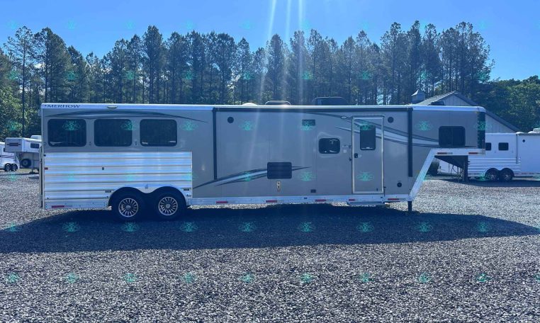 Side view of a long white and gray Merhow horse trailer parked on a gravel lot with tall pine trees and a blue sky behind it.