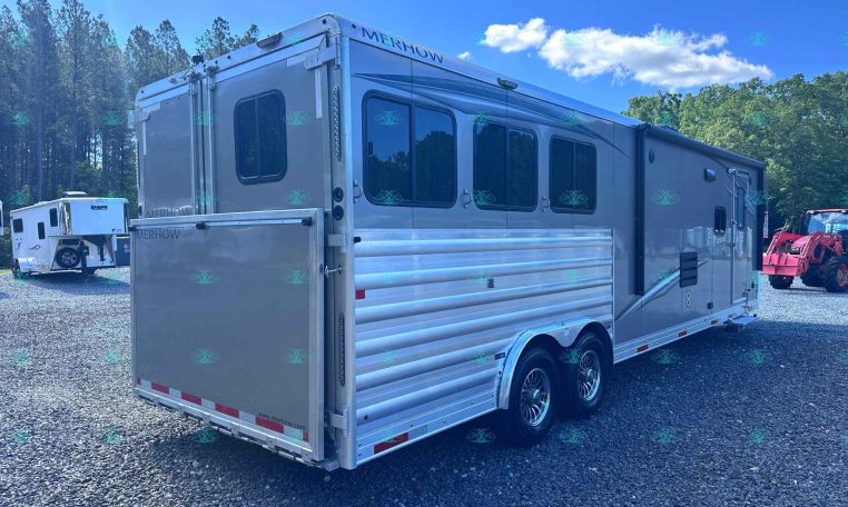 Silver Merhow horse trailer with multiple windows parked on a gravel lot, trees in the background.