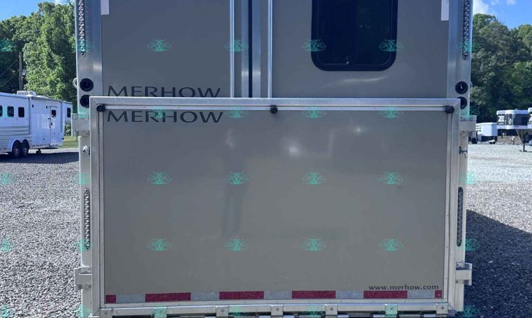 Rear view of a metal horse trailer with a small rounded window and dual doors, parked on gravel under a blue sky.