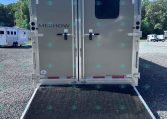 Back of a gray Merhow horse trailer with a lowered ramp, parked on a gravel lot under a blue sky.