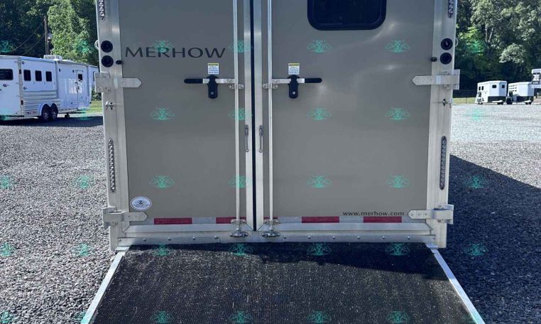 Back of a gray Merhow horse trailer with a lowered ramp, parked on a gravel lot under a blue sky.