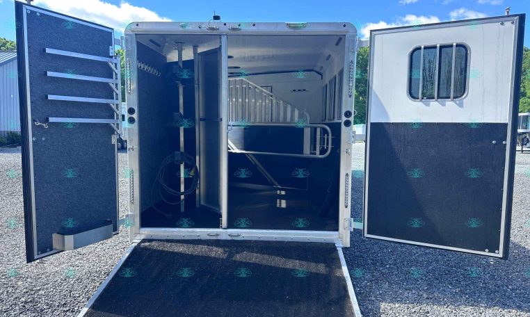 Open horse trailer interior with a divider, lowered ramp, and tack hooks along the left wall.