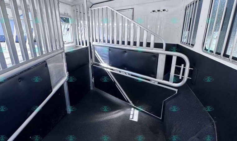 Interior of a metal horse trailer with divider stalls, gray rails, and black padded walls along the sides, ready for horses to ride.