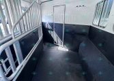 Interior of a white aluminum horse trailer with a center divider and entry door, black padded lower walls, and barred windows on the right.