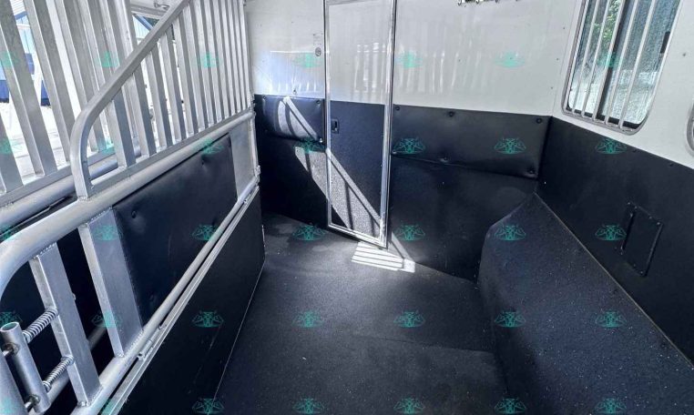 Interior of a white aluminum horse trailer with a center divider and entry door, black padded lower walls, and barred windows on the right.